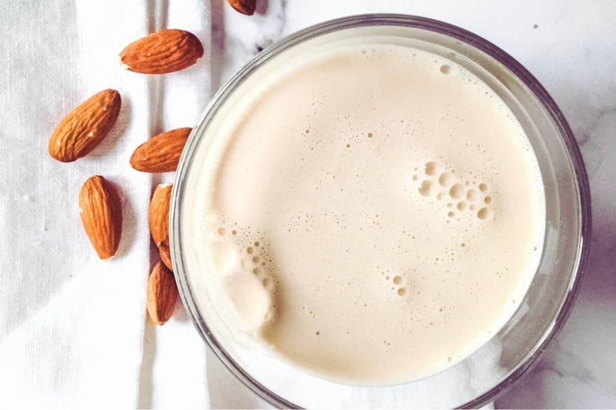 A glass of frothy almond milk and a few raw almonds on a kitchen towel.