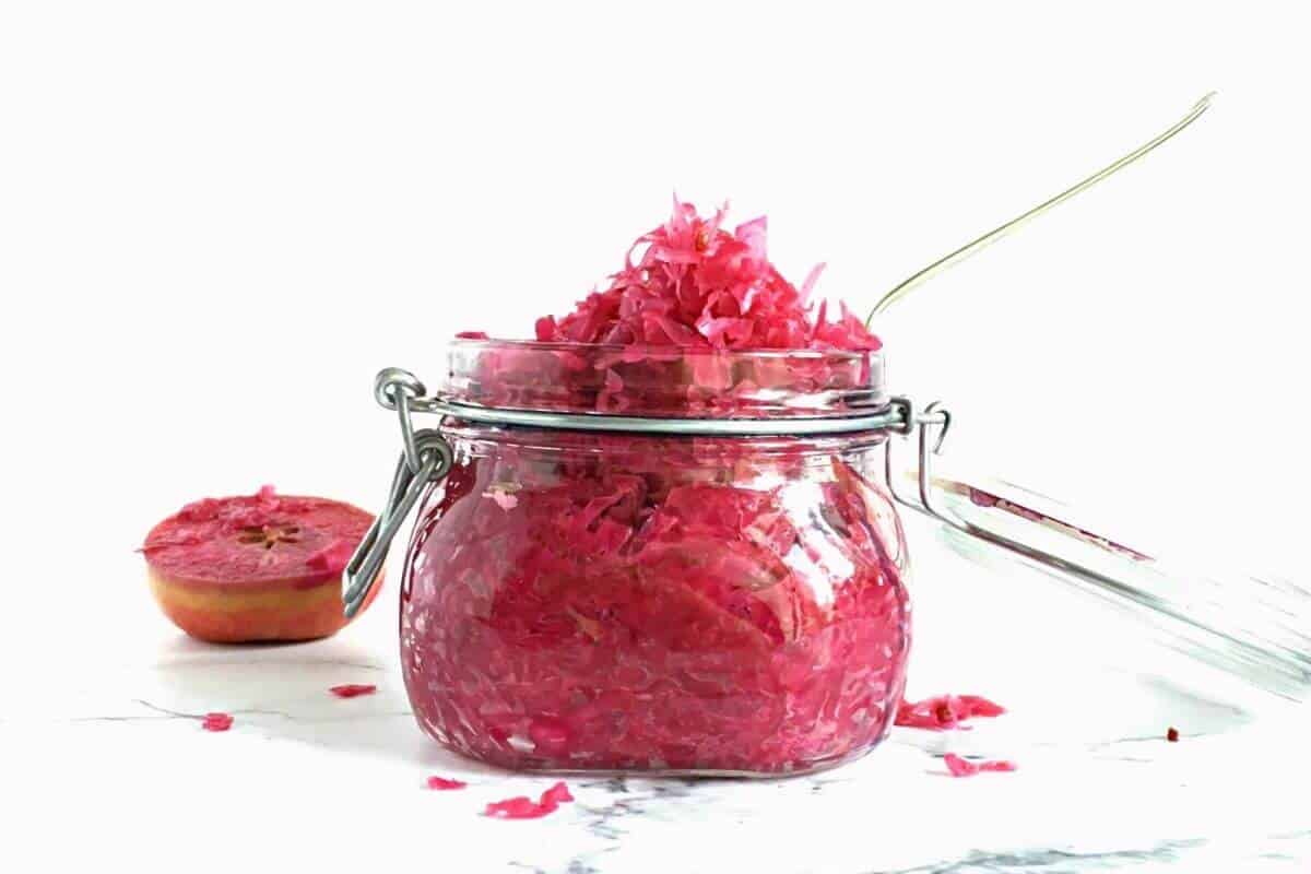 An opened mason jar filled with red cabbage sauerkraut sitting by an apple half.