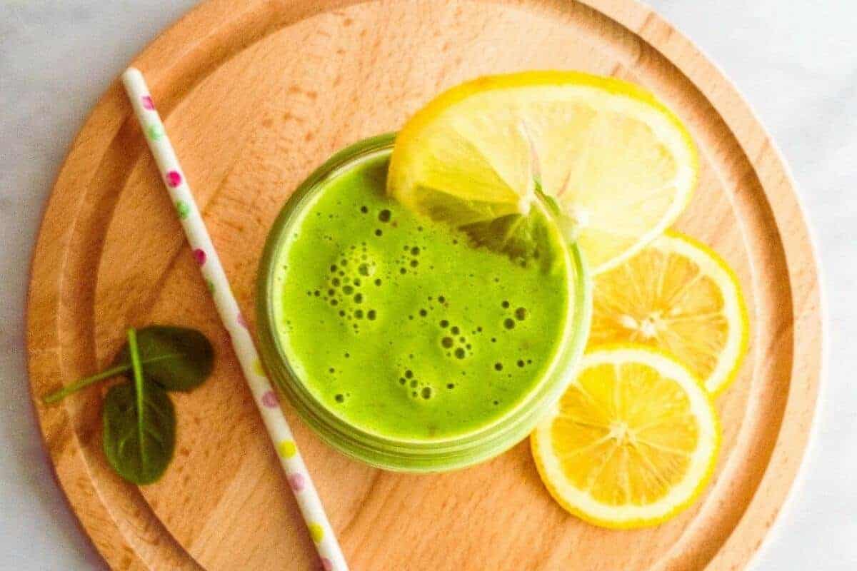 Glass of green smoothie on a chopping board with lemon slices and a straw.