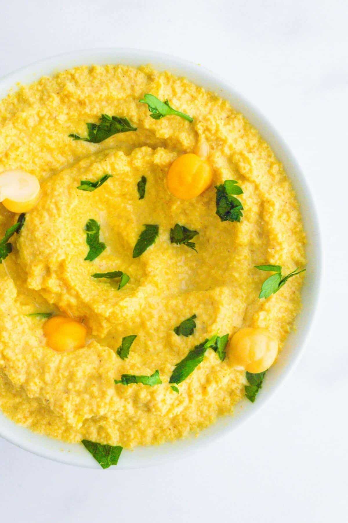A small bowl filled with yellow hummus, sprinkled with fresh herbs.