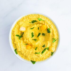 A bowl of bright yellow hummus with chickpeas and herbs.