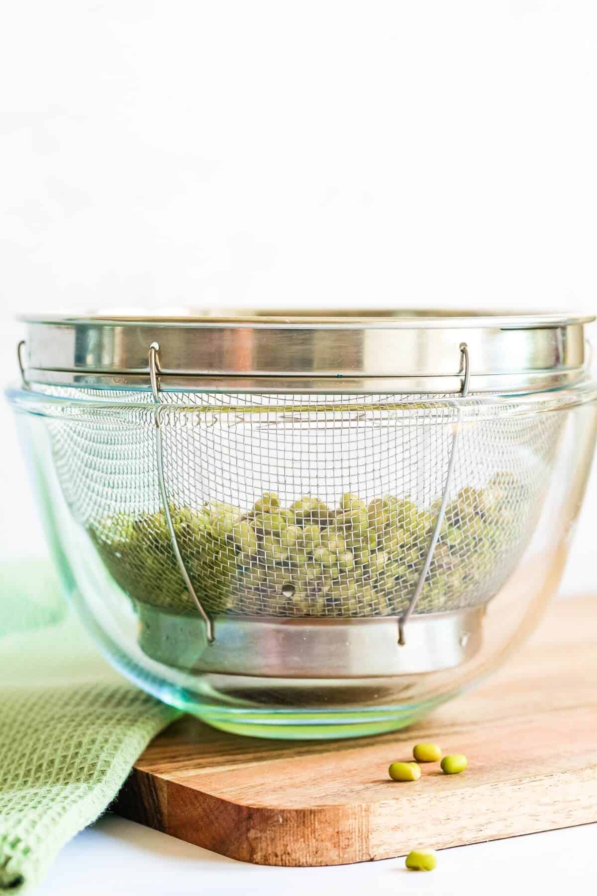 A metal strainer filled with mung beans is sat in a glass bowl on a wooden chopping board. A folded green tea towel is to the left.