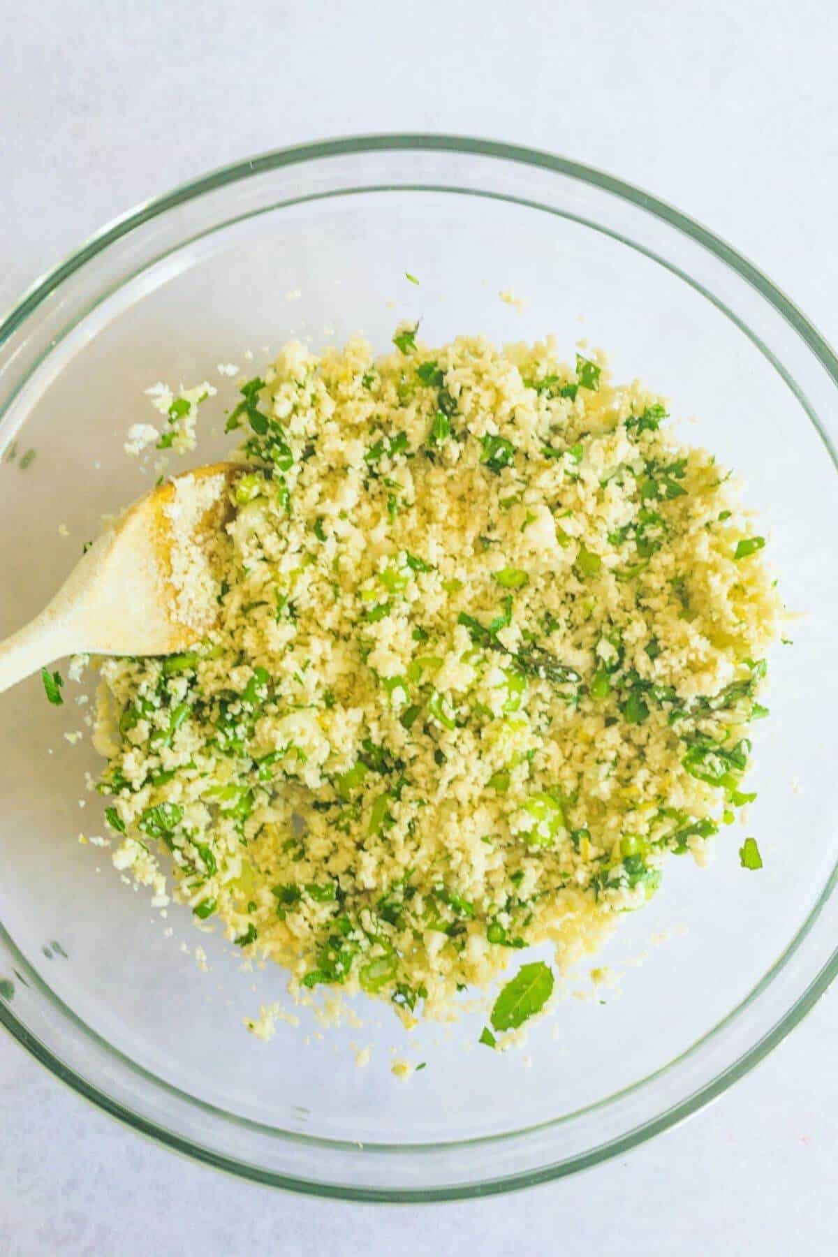 Green vegetables are stirred through a big glass bowl of cauliflower rice using a wooden spoon.