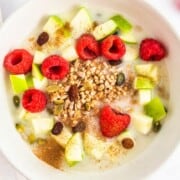 A bowl of cereal topped with fresh apple and raspberries.