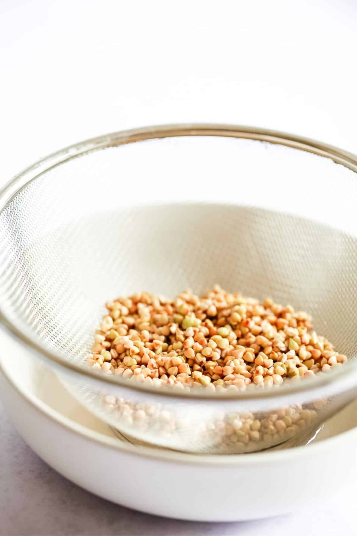 Raw buckwheat draining in a metal sieve placed over a bowl.