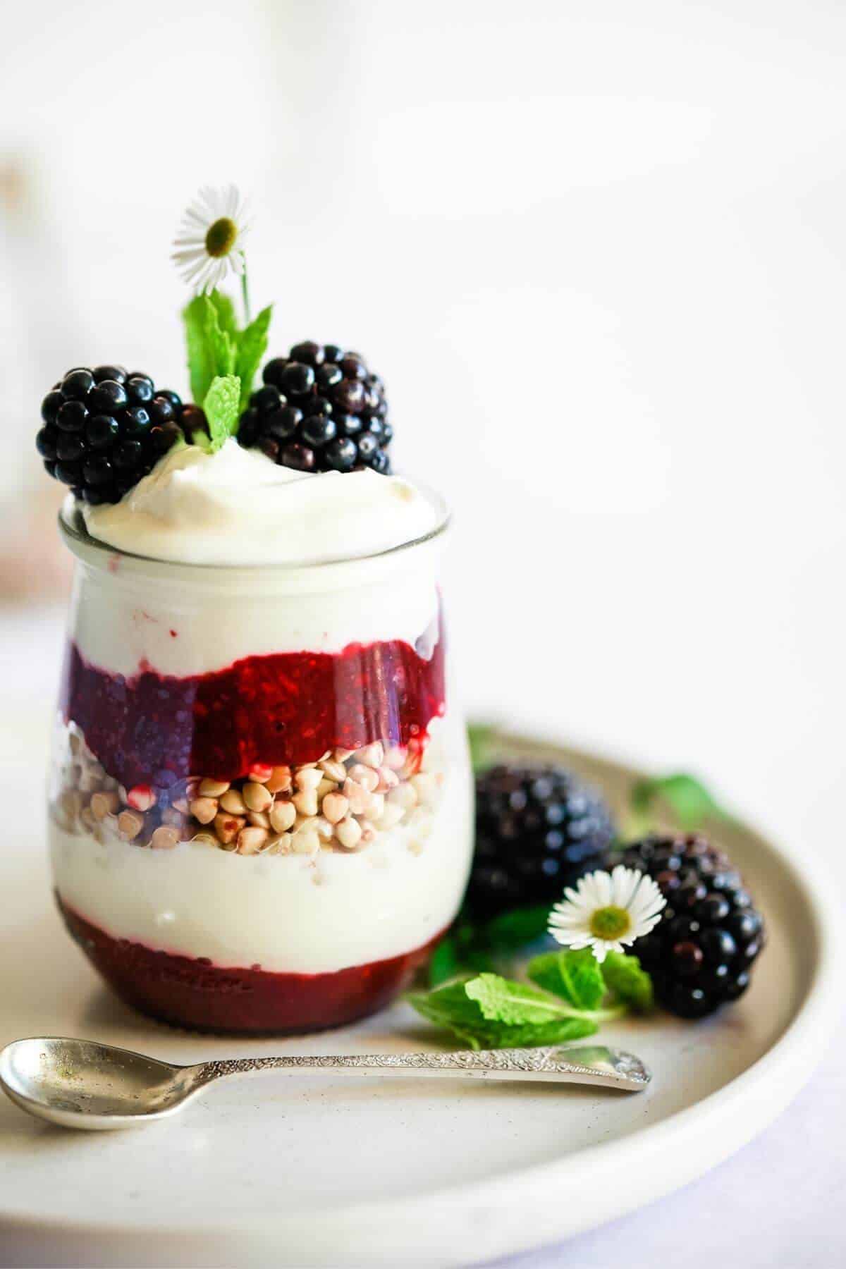 A blackberry parfait with a silver spoon sitting on a small plate garnished with mint leaves, daisies and fresh blackberries.