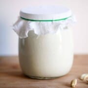 A jar full of yogurt, topped with cheesecloth sits on a wood surface. A couple of cashew nuts sit to the right.