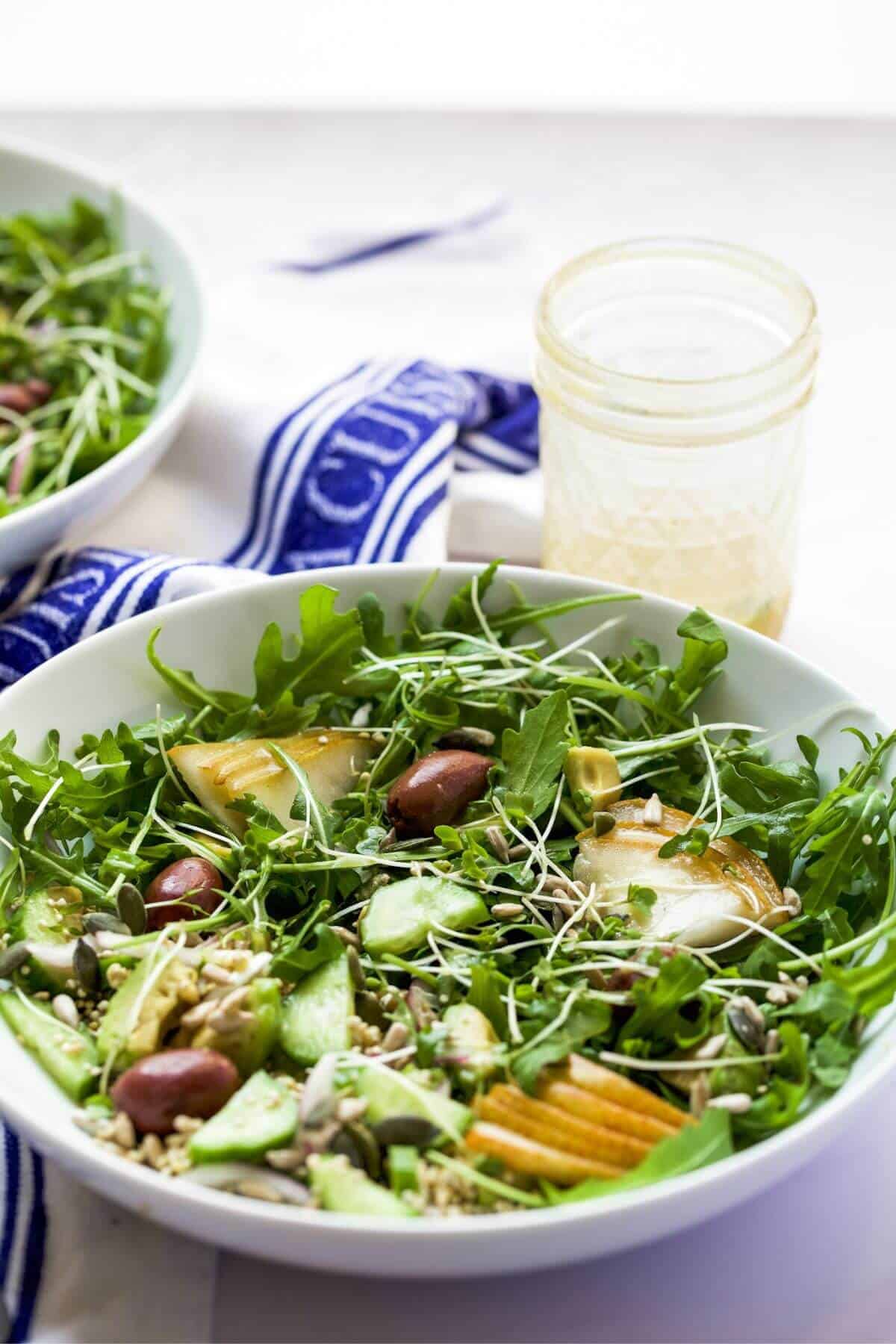 Two bowls on a table filled with a pear and rocket salad. A kitchen towel and small jar containing salad dressing by the bowls.