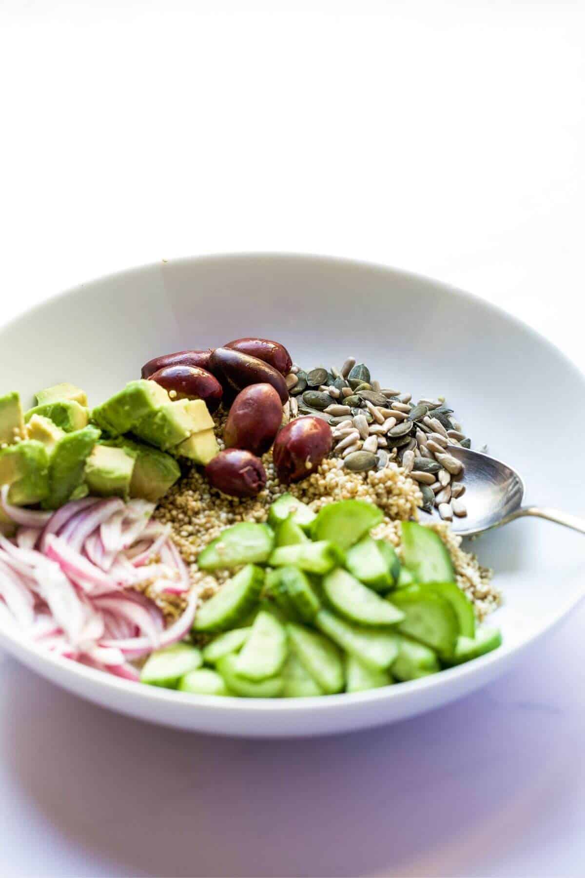 Chopped salad ingredients in a bowl including avocado, olives, seeds, cucumber, red onion and quinoa.