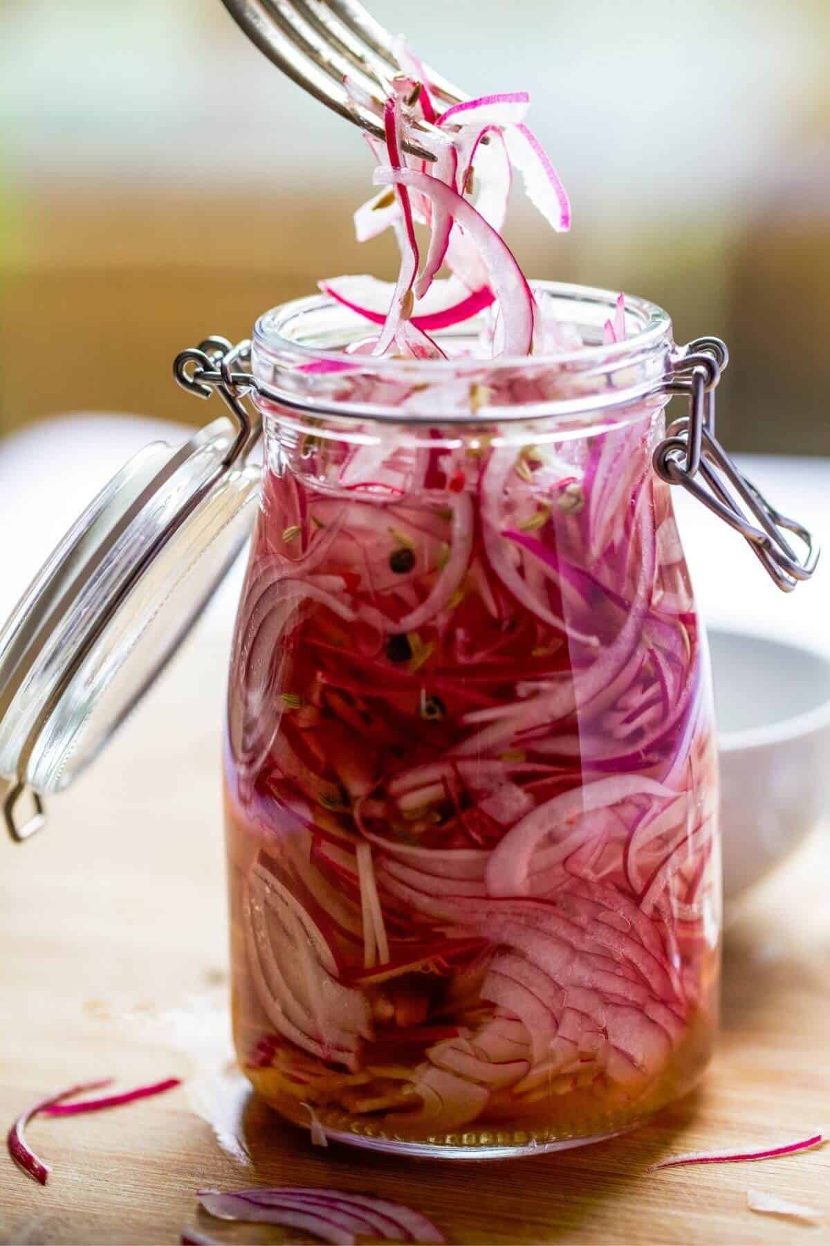 A silver fork lifts a few slices of red onion pickles from a small mason jar of pickles.