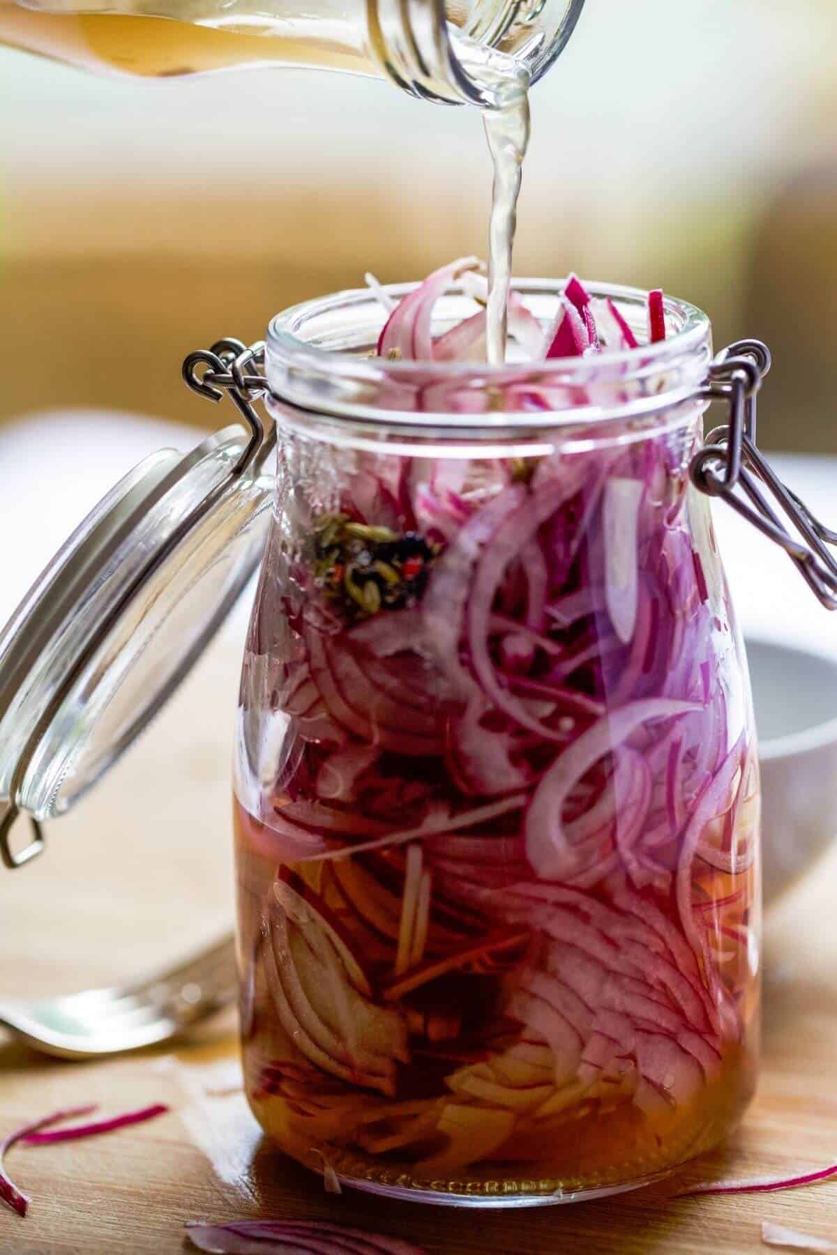 Vinegar is being poured into a mason jar filled with red onion slices.