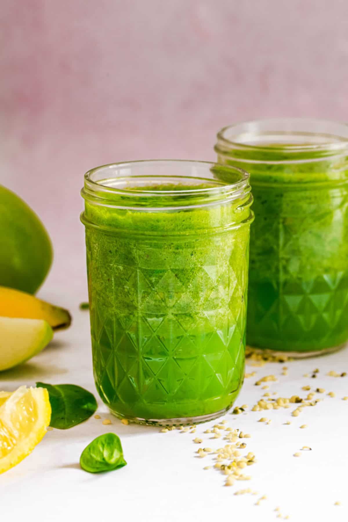 Two glasses of bright green smoothie with recipe ingredients nearby: sliced apple, banana, spinach, lemon slices & hemp seeds.