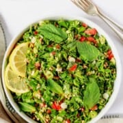 A big bowl of chopped parsley salad with fresh mint, tomatoes, buckwheat and topped with lemon slices. A kitchen towel and 2 forks nearby.
