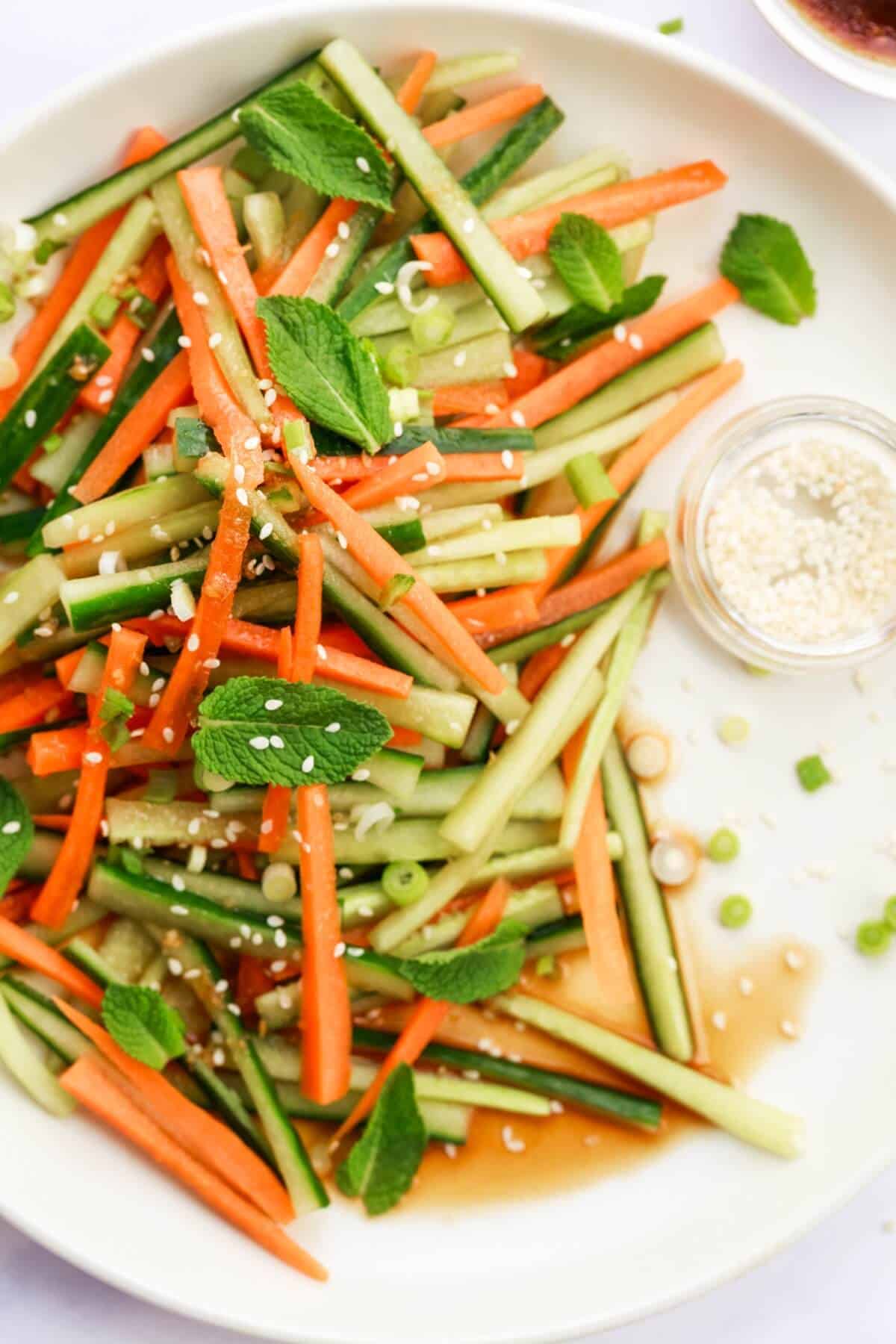 A plate of small carrot and cucumber sticks topped with fresh herbs, green onion and a tiny bowl with sesame seeds.