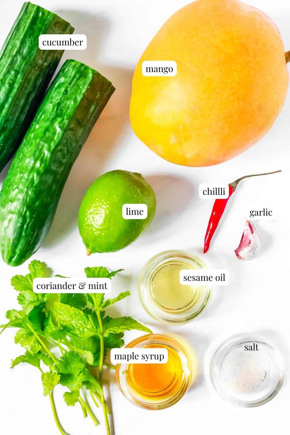 Labelled ingredients for a mango and cucumber salad laying on a white surface.