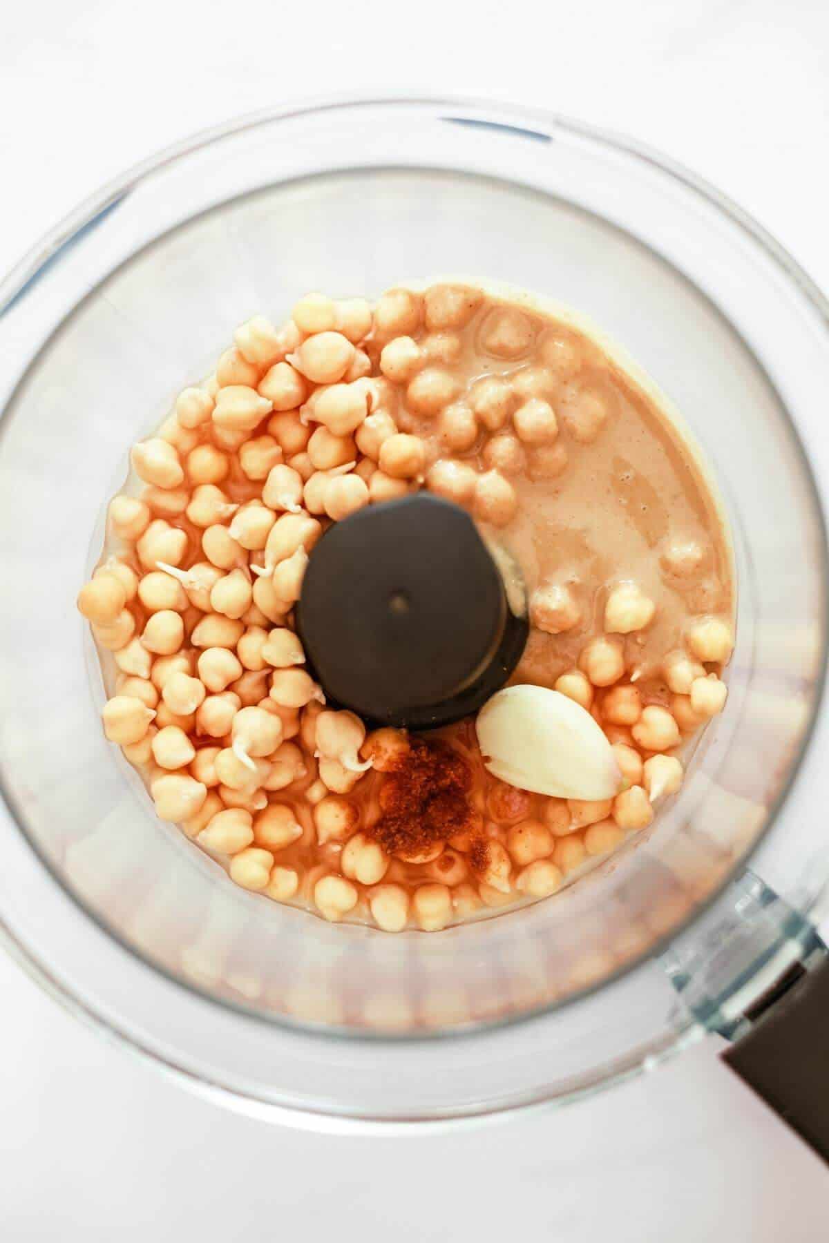 Top down view of a blender containing chickpeas, a garlic clove and some spice.