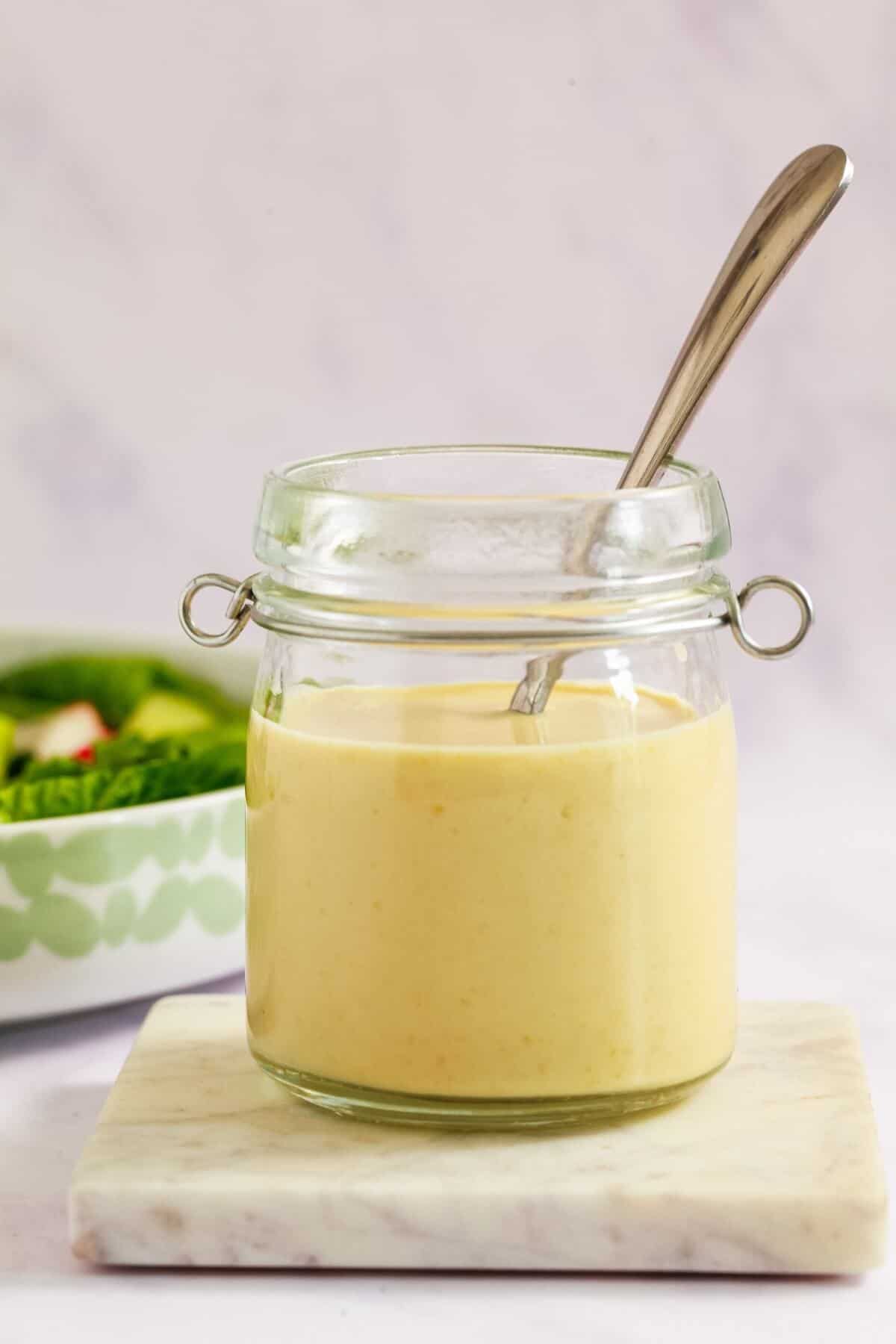 A open glass jar of salad dressing with a silver spoon sticking out of the top. The jar sits on a marble coaster with a salad bowl behind.
