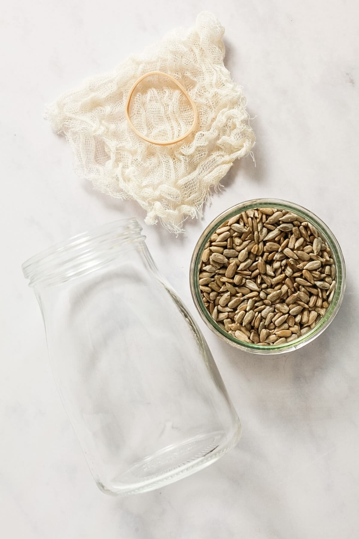 A small dish of sunflower seeds, glass jar, piece of cheesecloth and rubber band on a marble worksurface.