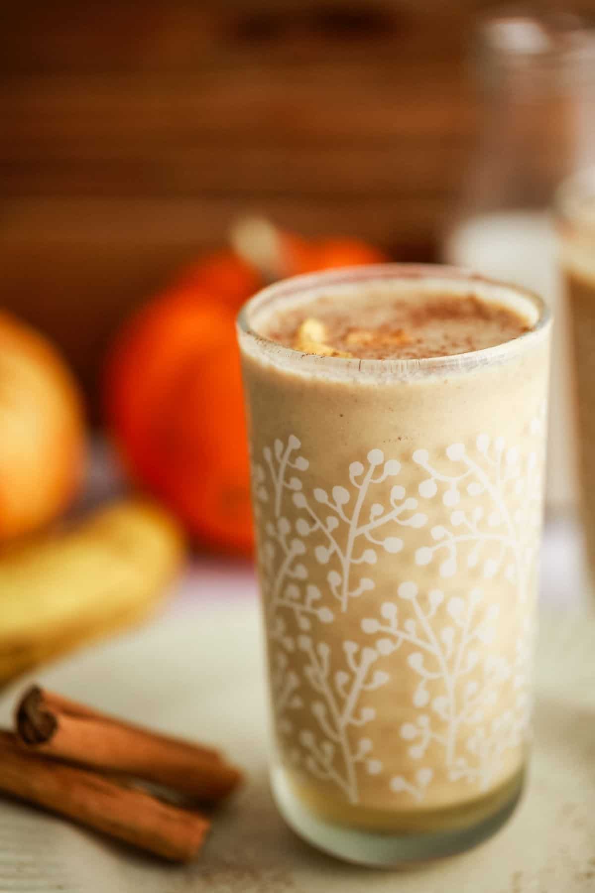 A tall glass of creamy drink topped with a little diced pumpkin. Sitting on a ceramic plate with cinnamon sticks. Pumpkins in the background.