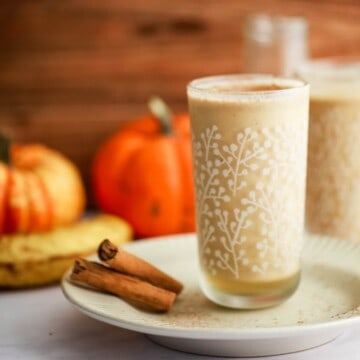 A tall glass of creamy drink on a small ceramic plate with cinnamon sticks, pumpkins and banana nearby.