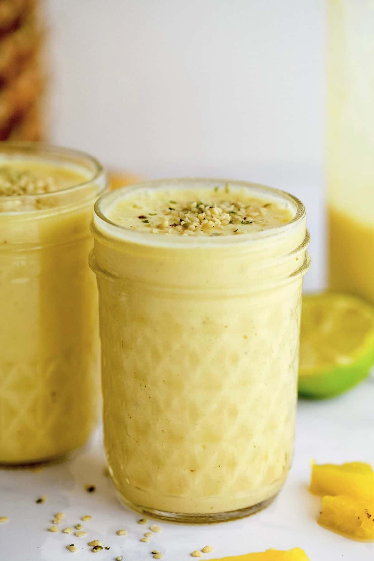 Two glass jars filled with pale yellow coloured drink topped with hemp seeds. Diced pineapple and half a lime by the jars.